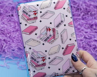 Puede incluir: Una funda de teléfono rosa y blanca con un patrón de pilas de libros con el texto "Tough Her & Die" y "Enemies to Lovers". Un cuchillo ensangrentado sobresale de la parte superior de la pila de libros.