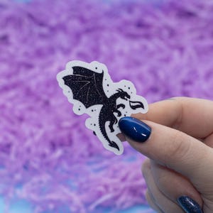 May include: A black glitter sticker of a dragon with its wings spread out. The dragon is silhouetted against a white background with small white stars.