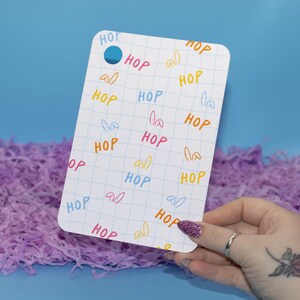 May include: A white rectangular card with rounded corners, featuring the word "HOP" in various colors and bunny ear illustrations. The card has a blue hole at the top and is held over purple shredded paper against a blue background.