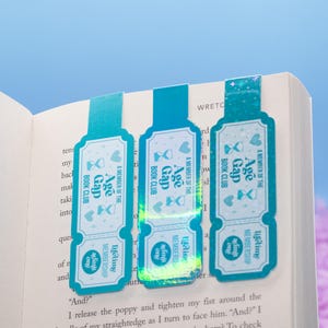 May include: Three teal and white bookmarks are inserted into a book. Each bookmark has a ticket design with the text "Age Gap Book Club" and "Lifetime Membership". The bookmarks have a gradient effect and are decorated with hearts and hourglasses.
