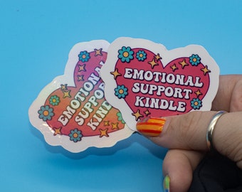 Puede incluir: Dos pegatinas en forma de corazón con acabado holográfico. Las pegatinas presentan el texto "Emotional Support Kindle" en una fuente retro con flores y estrellas de colores.