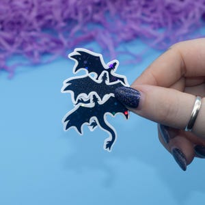 May include: A sticker featuring three black silhouette dragons with iridescent glitter accents. The dragons are layered on top of each other, creating a dynamic and whimsical design.