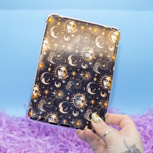 Peut inclure: Une coque de téléphone transparente avec un fond noir et un motif céleste doré. Le motif représente des soleils, des lunes et des étoiles avec des visages.