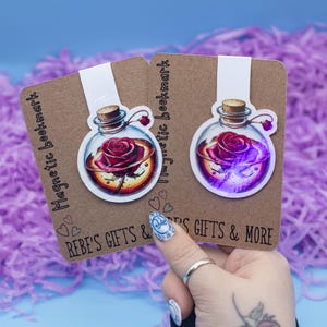 May include: A magnetic bookmark with a clear glass bottle containing a red rose. The bottle is surrounded by a gold-colored liquid. The bookmark is on a brown background with the text "Magnetic bookmark" and "Rebe's Gifts & More".