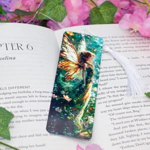 Puede incluir: Un marcapáginas con una ilustración colorida de un hada con alas iridiscentes, rodeada de follaje verde y flores. El hada lleva un vestido verde y tiene el pelo largo y suelto.