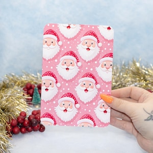 May include: A rectangular card featuring a repeating pattern of Santa Claus faces on a pink background with white polka dots. Santa is depicted with a red hat, white beard, and rosy cheeks. The card is held by a person with orange nail polish.