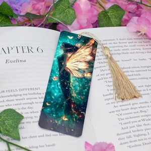 Puede incluir: Un marcapáginas con una ilustración de fantasía de un hada con alas brillantes y un vestido fluido. El marcapáginas tiene una borla y está colocado sobre una página de libro. La paleta de colores incluye verde azulado, naranja y negro.