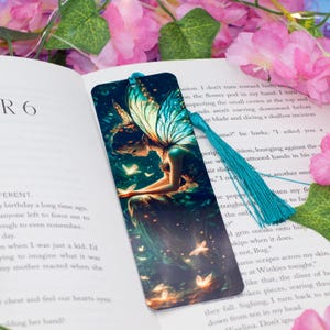 Puede incluir: Un marcapáginas azul verdoso con una ilustración colorida de un hada con alas iridiscentes y un vestido verde. El hada está rodeada de mariposas brillantes. El marcapáginas tiene un borla azul verdoso.