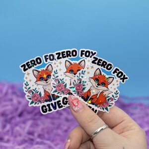 Puede incluir: Tres pegatinas holográficas con un zorro de dibujos animados y el texto "Zero Fox Given". Las pegatinas están sobre un fondo rosa y morado.