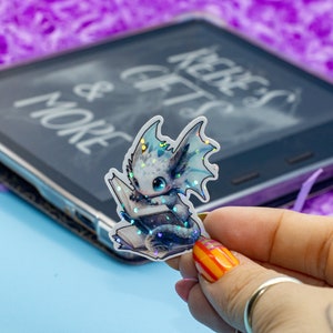 May include: A holographic sticker of a blue and white dragon reading a book. The dragon has iridescent scales and a book with a gold spine.