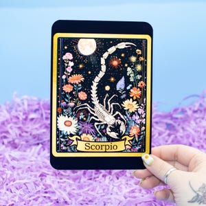 May include: A rectangular black card with a gold border features a detailed illustration of a scorpion skeleton surrounded by colorful flowers and a full moon. The word "Scorpio" is written on a banner at the bottom.