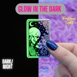 May include: A sticker featuring two skeletons kissing, surrounded by flowers. The sticker glows in the dark, and the glow is shown on the left side of the image. The right side of the image shows the sticker in daylight.