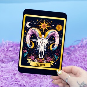 Può includere: Una carta rettangolare nera con bordo dorato raffigurante un'illustrazione di un teschio con corna colorate, circondato da stelle, fiori e la scritta "Aries" su uno stendardo. La carta è tenuta su uno sfondo viola e blu.