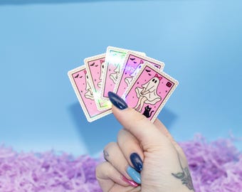 Può includere: Una mano tiene in mano una pila di adesivi rosa con un'illustrazione di un fantasma. Ogni adesivo presenta un fantasma bianco con un fiocco rosa, un gatto nero e un pipistrello. Gli adesivi hanno un design a francobollo con un bordo ondulato. Lo sfondo è blu e viola.