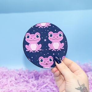 May include: A circular coaster with a navy blue background, patterned with pink frogs and white stars. The frogs have large eyes and are smiling. The coaster is held by a hand with a dark blue nail.