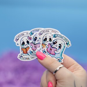 Puede incluir: Tres pegatinas holográficas iridiscentes con un fantasma de dibujos animados llorando por personajes ficticios. Cada pegatina tiene el texto "Crying over fictional characters" y un fantasma sosteniendo un libro.