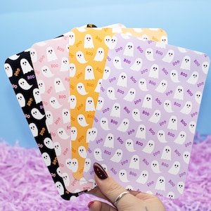 May include: A hand displays a selection of Halloween-themed cards. Each card has a repeating pattern of white ghosts and the word "BOO" in various colours. The cards are black, pink, yellow, and purple.