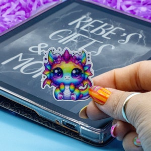 May include: A rainbow-coloured dragon sticker with a cute expression. The sticker is on a black tablet with the text "Rebe's Gifts & More" on the screen.