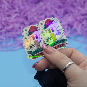 Op de afbeelding: Twee holografische stickers met een cartoongeest met een kerstmuts en een lichtsnoer. De stickers hebben de tekst "Sorta Merry, Sorta Scary". De stickers staan op een blauwe achtergrond.