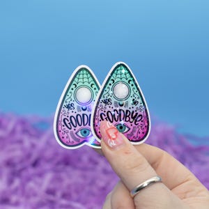 May include: Two holographic pink and green ouija board shaped stickers with the words "Goodbye" and "Good" on them. The stickers are held by a hand with a ring on the finger.