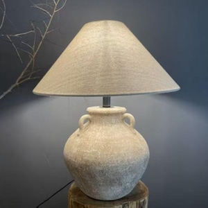 Puede incluir: Lámpara de mesa beige con pantalla cónica. La base de la lámpara es un diseño en forma de urna con dos pequeñas asas. La lámpara está colocada sobre un tocón de madera, con un fondo oscuro.