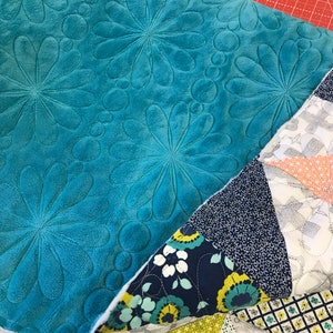 May include: A teal blue quilted fabric with a floral pattern. The fabric is folded over a patchwork quilt with various patterns in shades of blue, green, yellow, and white.