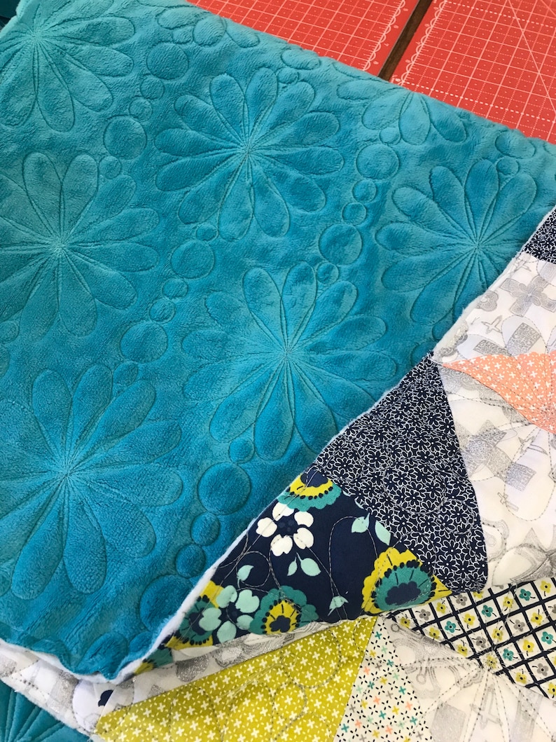 May include: A teal blue quilted fabric with a floral pattern. The fabric is folded over a patchwork quilt with various patterns in shades of blue, green, yellow, and white.