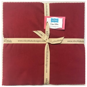May include: A red fabric square wrapped in a beige ribbon with a label that reads "Riley Blake Cotton Solids RED Designers" and the website address "www.rileyblakedesigns.com".