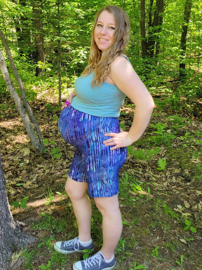 Puede incluir: Una mujer embarazada con un top azul claro sin mangas y pantalones cortos de rayas azules y moradas. Est&aacute; de pie en un bosque con &aacute;rboles y hojas verdes.
