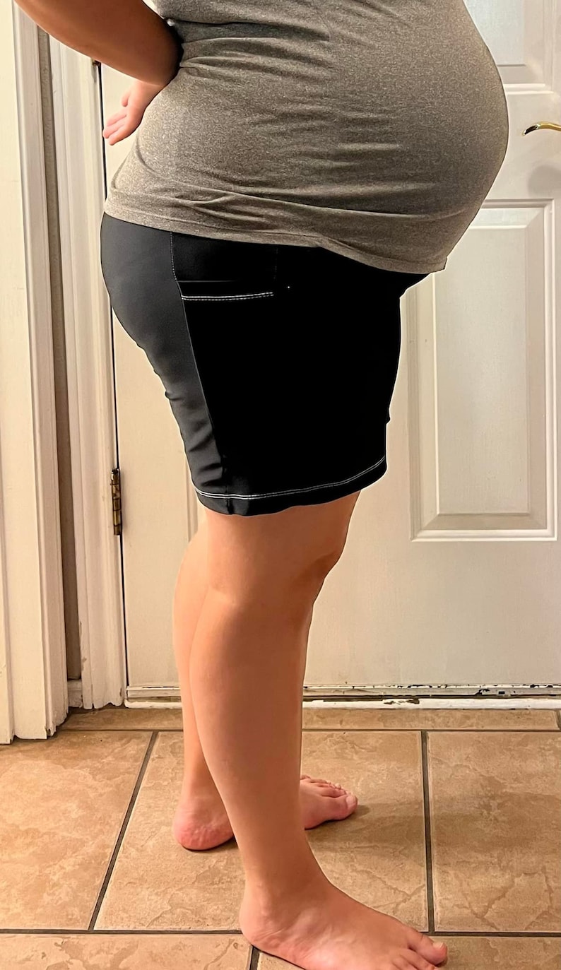 Puede incluir: Una mujer con una camiseta de maternidad gris y pantalones cortos de maternidad negros con costuras blancas. Los pantalones cortos tienen un bolsillo lateral.