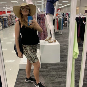 Puede incluir: Una mujer con un top negro de tirantes, una falda estampada en marr&oacute;n y blanco y zapatillas negras. Lleva un sombrero de paja de ala ancha y gafas de sol. Est&aacute; de pie frente a un espejo en una tienda de ropa.
