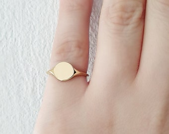 May include: A gold signet ring with a smooth, oval-shaped top. The ring is worn on the middle finger of a hand with nude nail polish.