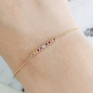 Op de afbeelding: Een gouden kettingarmband met een rij kleurrijke edelstenen. De edelstenen zijn gerangschikt in een verloop, beginnend met oranje en overgaand naar rood.
