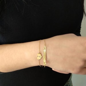 Op de afbeelding: Twee gouden armbanden om een pols. De ene armband heeft een hartvormige bedel met de letter A erop gegraveerd. De andere armband heeft een staafvormige bedel met een geometrisch ontwerp.