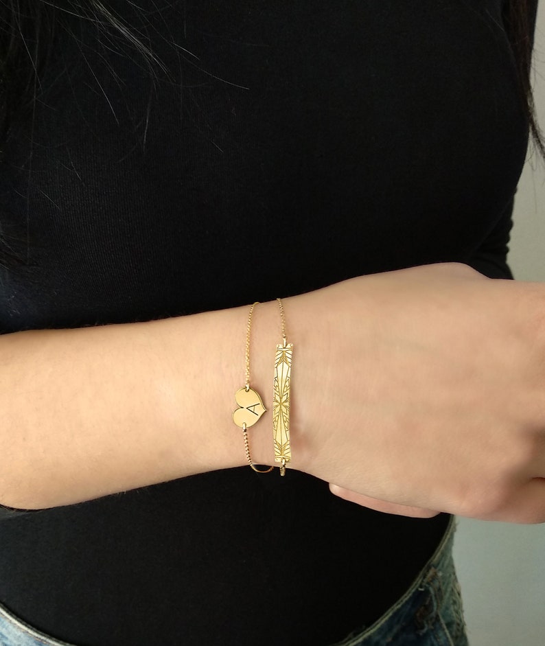 Op de afbeelding: Twee gouden armbanden om een pols. De ene armband heeft een hartvormige bedel met de letter A erop gegraveerd. De andere armband heeft een staafvormige bedel met een geometrisch ontwerp.