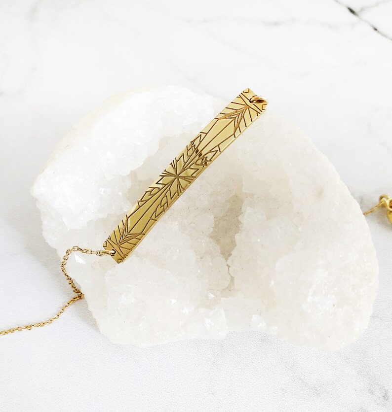 Op de afbeelding: Een gouden ketting met een delicate ketting en een bar hanger met een geometrisch sneeuwvlokkenpatroon.