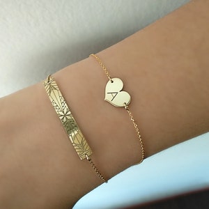 Op de afbeelding: Twee gouden armbanden om een pols. De ene armband heeft een hartvormige bedel met de letter A erop gegraveerd. De andere armband heeft een staafvormige bedel met een geometrisch ontwerp.