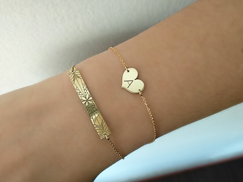 Op de afbeelding: Twee gouden armbanden om een pols. De ene armband heeft een hartvormige bedel met de letter A erop gegraveerd. De andere armband heeft een staafvormige bedel met een geometrisch ontwerp.