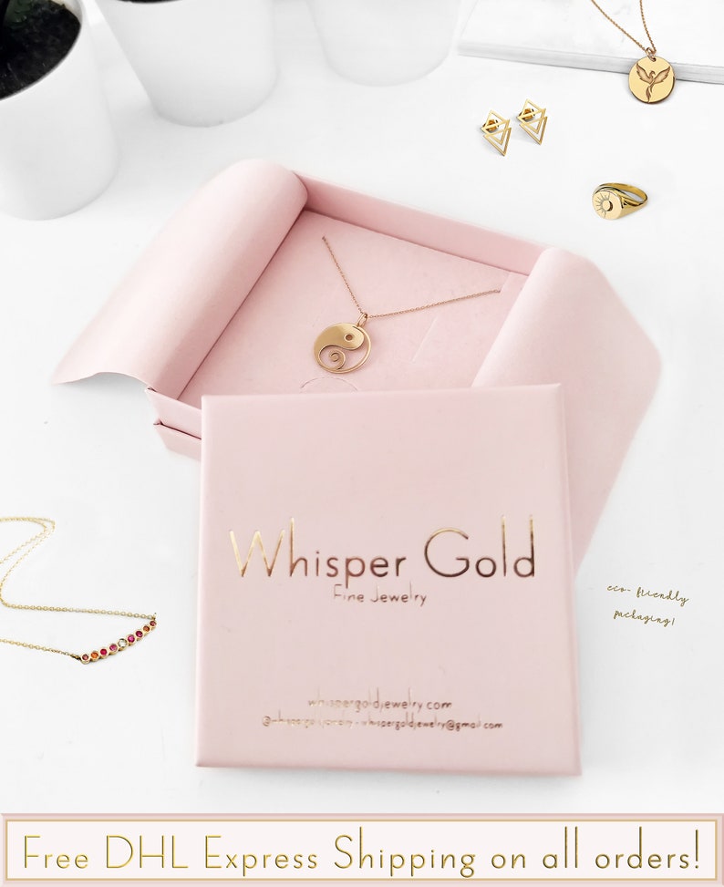 May include: A pink jewelry box with the text "Whisper Gold Fine Jewelry" and the website address "whispergoldjewelry.com". Inside the box is a gold necklace with a yin yang pendant.