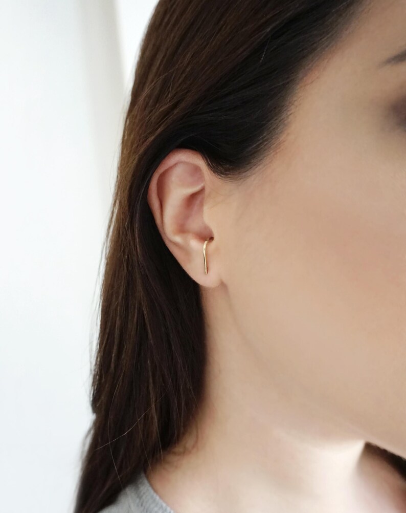 May include: A close-up of a person's ear with a thin gold bar earring. The earring is worn in the upper cartilage of the ear.