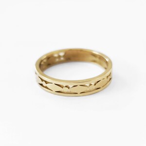 May include: A gold-colored ring with a repeating leaf-like pattern around the band. The ring is set against a white background, highlighting its intricate design. The ring's band is approximately 0.2 inches wide.
