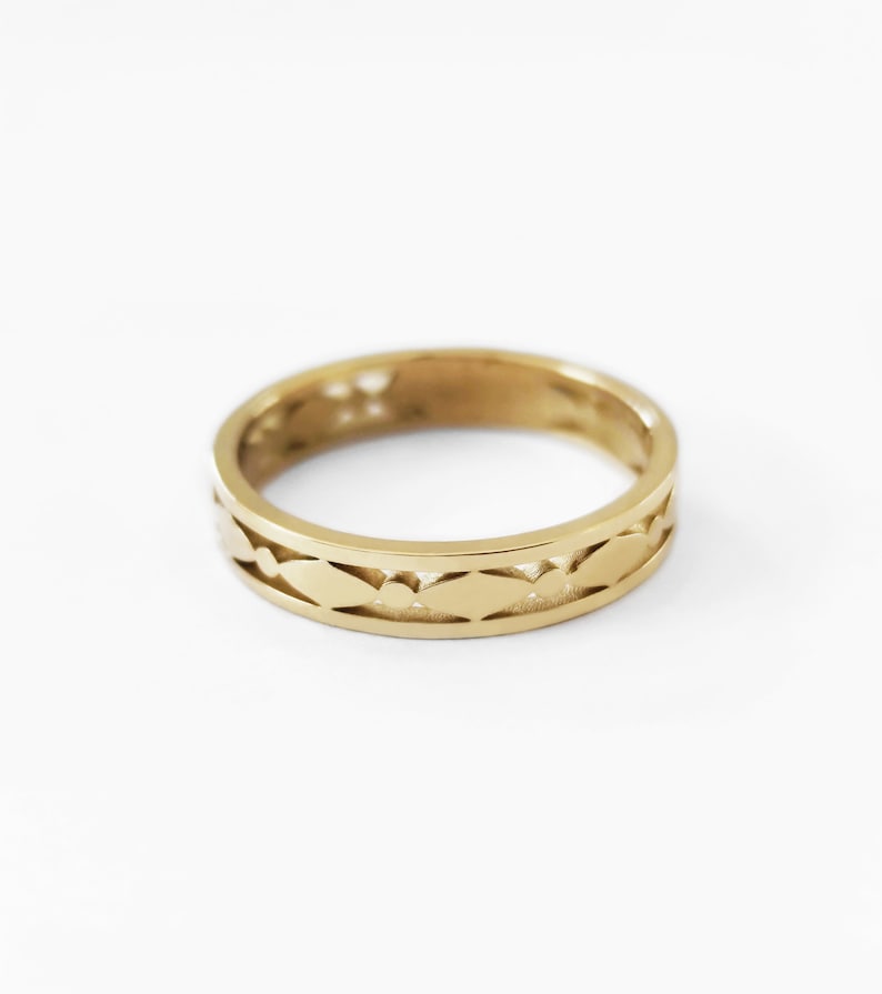 May include: A gold-colored ring with a repeating leaf-like pattern around the band. The ring is set against a white background, highlighting its intricate design. The ring's band is approximately 0.2 inches wide.