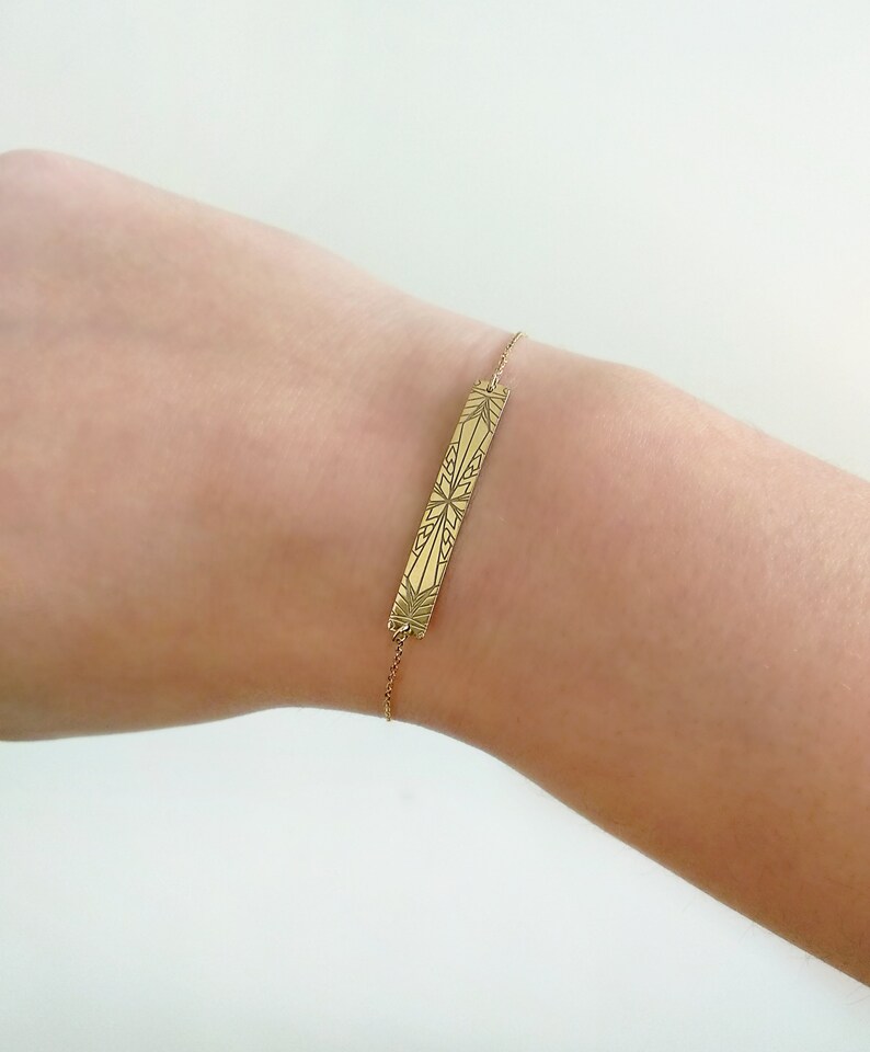 Op de afbeelding: Een gouden armband met een delicate ketting en een rechthoekige bedel met een geometrisch ontwerp.
