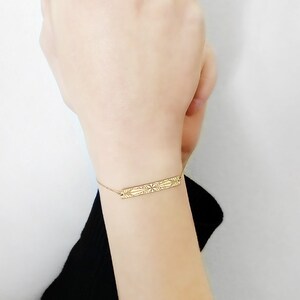 Op de afbeelding: Een gouden armband met een delicate ketting en een rechthoekige bedel met een ingewikkeld geometrisch ontwerp.