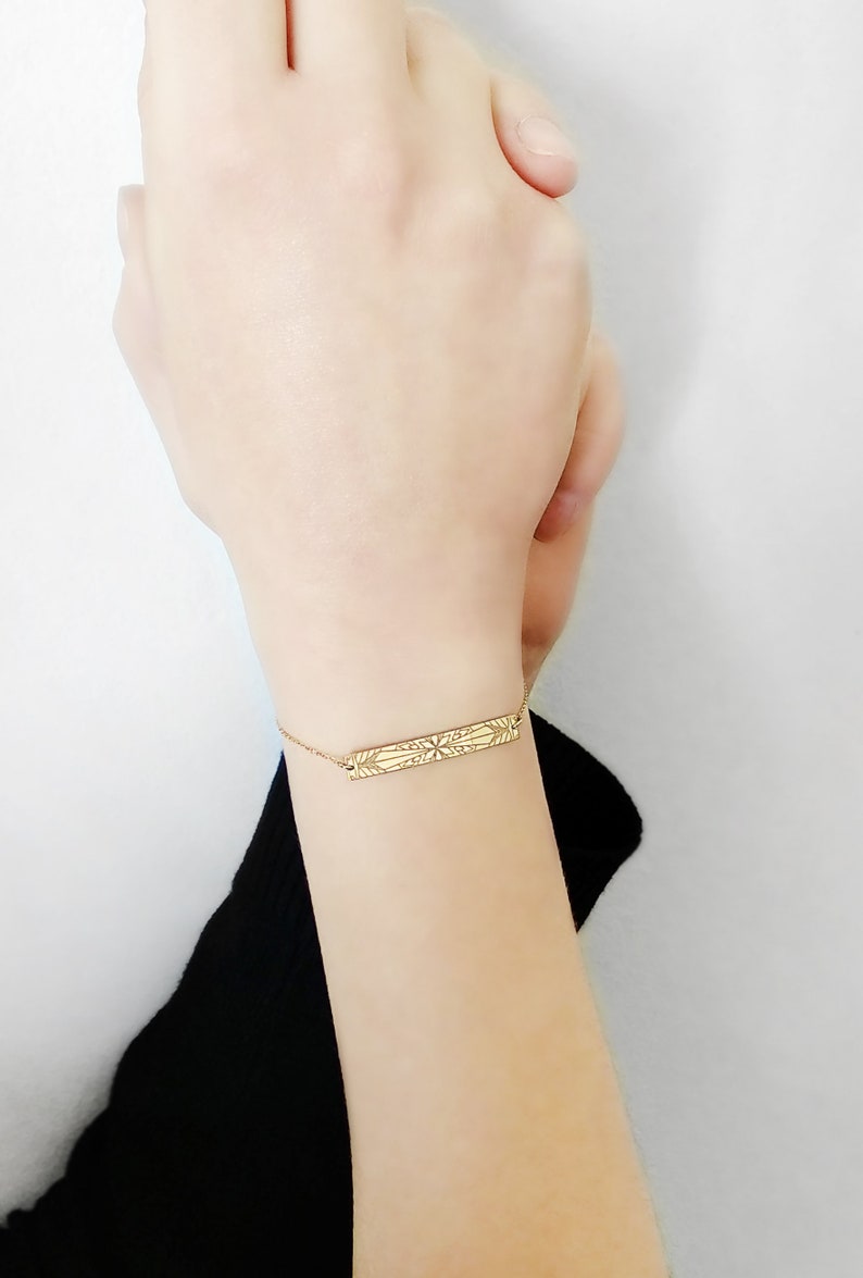 Op de afbeelding: Een gouden armband met een delicate ketting en een rechthoekige bedel met een ingewikkeld geometrisch ontwerp.
