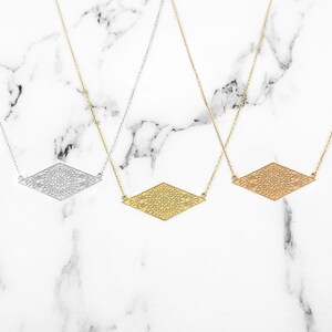May include: Three delicate necklaces with a geometric diamond-shaped pendant featuring a detailed, intricate design. The necklaces are shown on a white marble background. The pendants are silver, gold, and rose gold.
