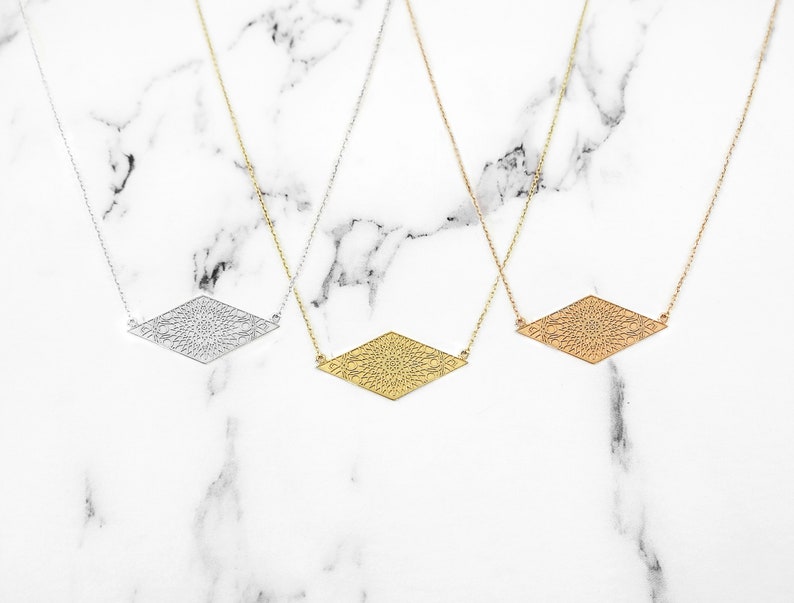 May include: Three delicate necklaces with a geometric diamond-shaped pendant featuring a detailed, intricate design. The necklaces are shown on a white marble background. The pendants are silver, gold, and rose gold.