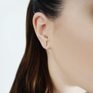 May include: A close-up of a person's ear with a gold, curved, minimalist earring. The earring is a simple, thin line that curves slightly.