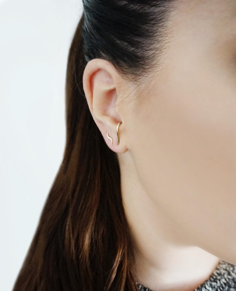May include: A close-up of a person's ear with a gold, curved, minimalist earring. The earring is a simple, thin line that curves slightly.