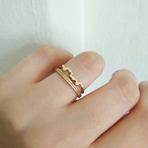 May include: Two gold rings on a finger. The top ring has a wavy design and the bottom ring is a simple band.